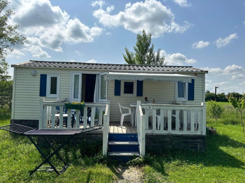 Mobil-home Sun Living avec terrasse pour vacances au camping Mobil-home Sun Living numéro 36 avec terrasse en bois, chaises blanches et auvent sous un ciel nuageux.